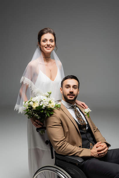 Mariée avec bouquet et marié arabe en fauteuil roulant souriant à la caméra isolée sur gris  - Photo, image