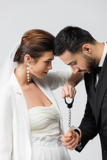 Невеста, держащая наручники рядом с расстроенным мусульманским женихом, изолированным на сером  - Фото, изображение