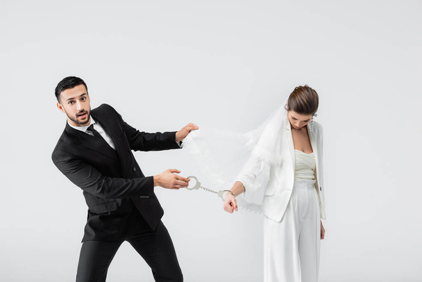 Muslimischer Bräutigam zieht Handschellen und Schleier trauriger Braut isoliert auf grau  - Foto, Bild