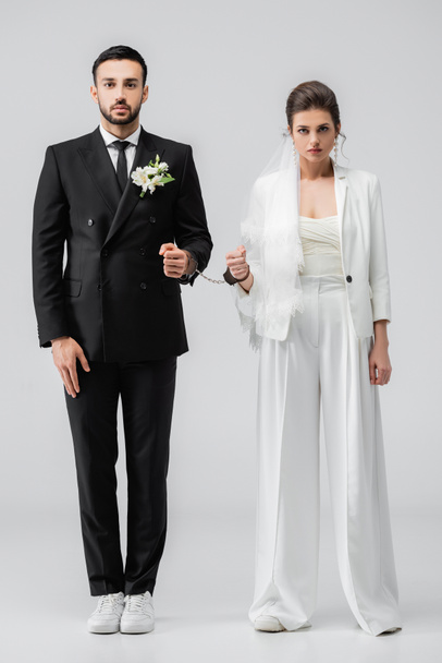Interrassische Bräutigam und Braut in Hochzeitskleidung und Handschellen Blick in die Kamera auf weißem Hintergrund - Foto, Bild