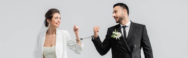 Smiling bride in handcuffs looking at cheerful muslim groom isolated on grey, banner  - Photo, Image