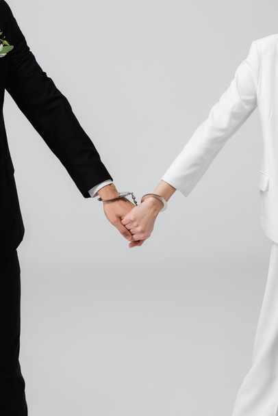 Cropped view of bride and groom in handcuffs holding hands isolated on grey  - Photo, Image