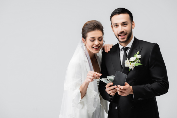 Noiva sorridente olhando para a carteira com dinheiro em mãos de noivo muçulmano isolado em cinza  - Foto, Imagem