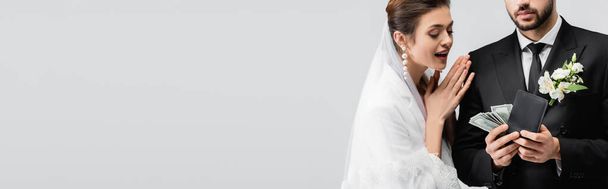 Usmívající se nevěsta dívá na peníze v rukou ženicha izolovaných na šedé, prapor  - Fotografie, Obrázek
