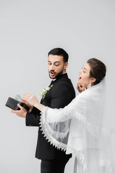 Ενθουσιασμένη νύφη στέκεται κοντά στο αραβικό γαμπρό με δολάρια και πορτοφόλι απομονωμένα στο γκρι  - Φωτογραφία, εικόνα