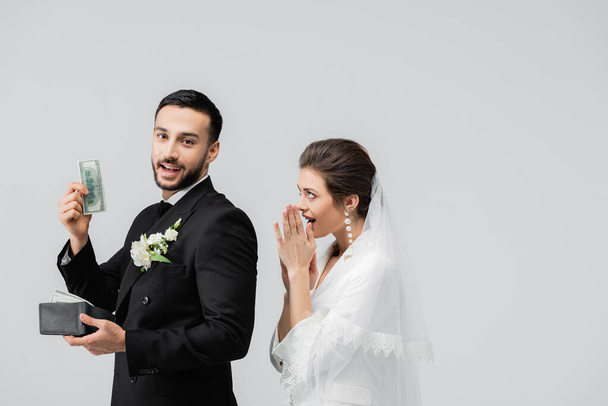 Souriant marié arabe tenant de l'argent près de mariée excitée isolé sur gris  - Photo, image