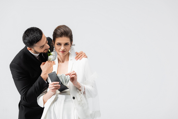 Μουσουλμάνος γαμπρός αγκαλιάζει τη νύφη με μετρητά και πορτοφόλι απομονωμένο σε γκρι  - Φωτογραφία, εικόνα