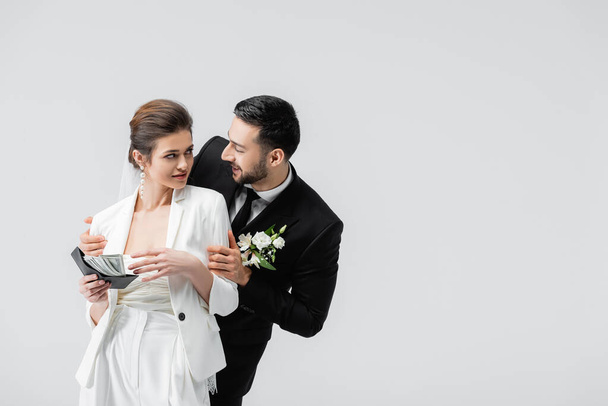 Sorridente sposo arabo abbracciare sposa con contanti e portafoglio isolato su grigio  - Foto, immagini