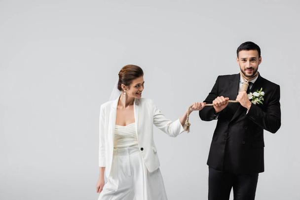 Χαμογελώντας νύφη τραβώντας σχοινί γύρω από το λαιμό του αραβικού φίλου απομονωμένο σε γκρι  - Φωτογραφία, εικόνα