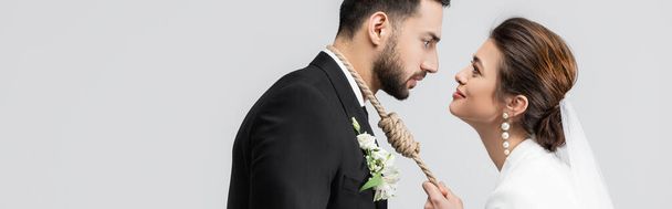 Seitenansicht der jungen Braut in Schleier mit Slip-Knoten am Hals des arabischen Bräutigams isoliert auf grau, Banner  - Foto, Bild