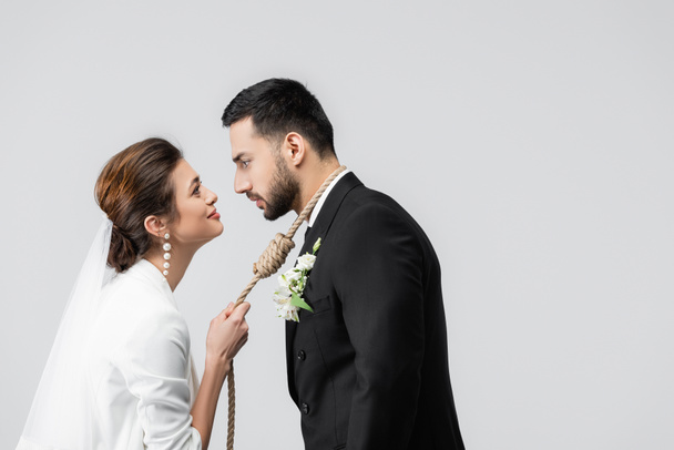 Vue latérale de la mariée souriante tenant le noeud glissant sur le cou du marié musulman isolé sur gris  - Photo, image