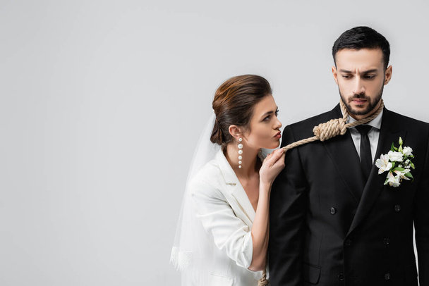 Νύφη κρατώντας κόμπο ολίσθησης στο λαιμό του λυπημένου αραβικού γαμπρού απομονωμένη σε γκρι  - Φωτογραφία, εικόνα