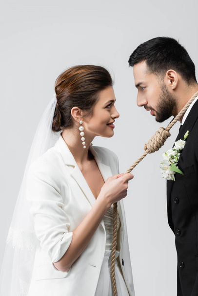 Side view of smiling bride pulling rope around neck of muslim groom isolated on grey  - Photo, Image