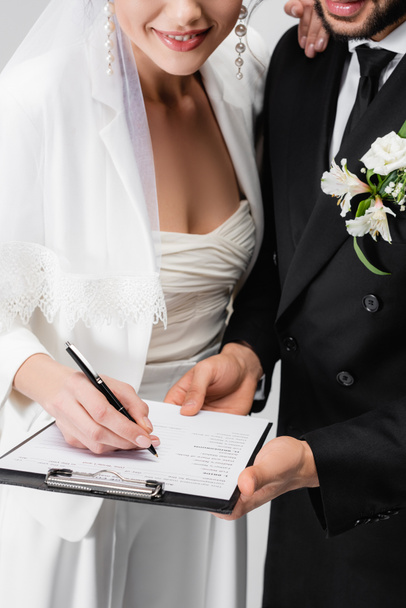 Обрезанный вид улыбающейся невесты, подписывающей свадебный контракт рядом с женихом, изолированным на сером  - Фото, изображение