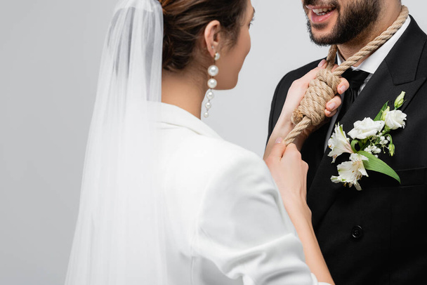 Νεαρή νύφη φορώντας κόμπο ολίσθησης στο λαιμό του γαμπρού απομονωμένη σε γκρι  - Φωτογραφία, εικόνα