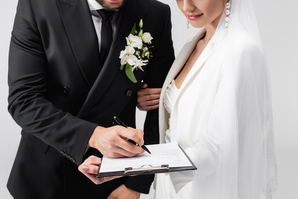 Vista cortada do contrato de assinatura do noivo perto da noiva em véu isolado em cinza  - Foto, Imagem