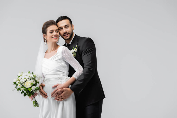 Glimlachende zwangere bruid staat naast moslim bruidegom geïsoleerd op grijs  - Foto, afbeelding
