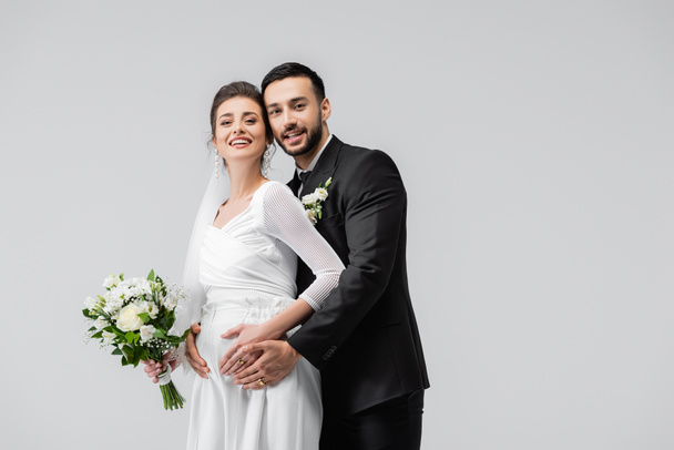 Joven novio musulmán abrazando alegre novia embarazada con ramo de boda aislado en gris  - Foto, imagen