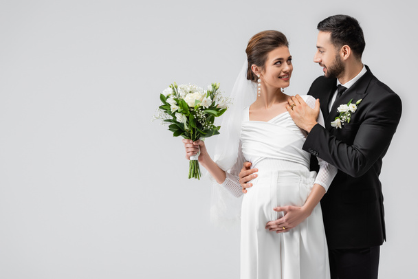 Junger muslimischer Mann umarmt fröhliche schwangere Braut isoliert auf grau  - Foto, Bild