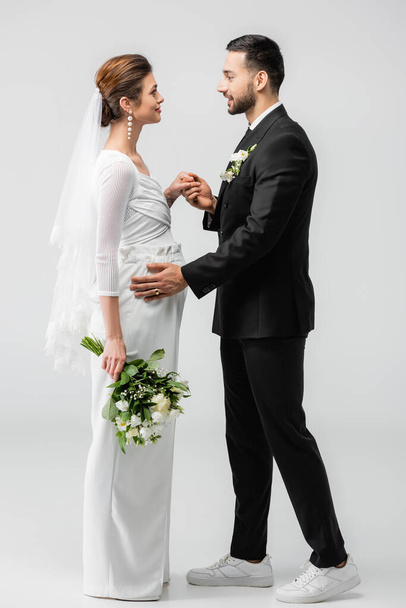 Vista lateral del novio musulmán cogido de la mano de la novia embarazada sobre fondo gris  - Foto, Imagen
