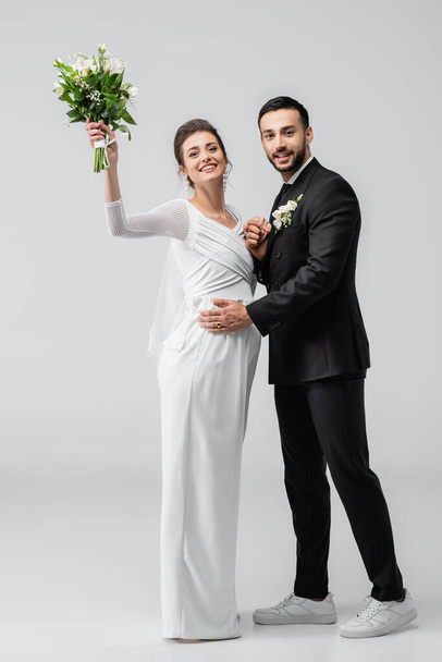 Fröhlich schwangere Braut hält Blumenstrauß neben muslimischem Bräutigam auf grauem Hintergrund  - Foto, Bild