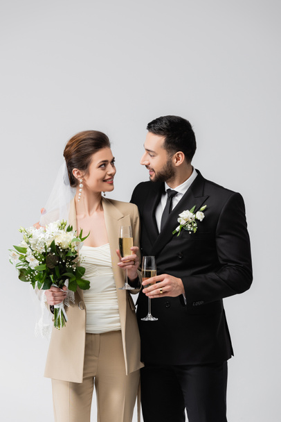 Улыбающаяся невеста, держащая букет и бокал шампанского рядом с мусульманским женихом, изолированным на сером  - Фото, изображение