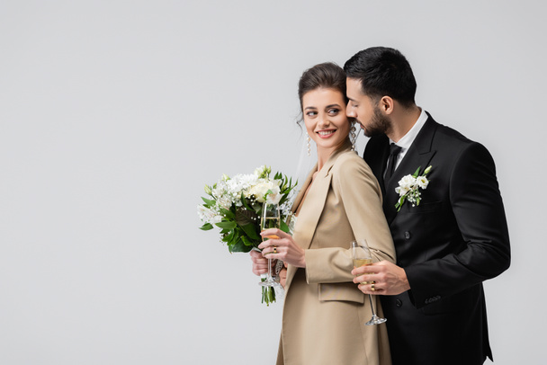 Novia sonriente con ramo de bodas mirando al novio árabe con champán aislado en gris  - Foto, imagen