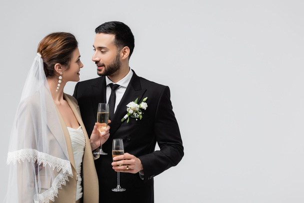 Lächelndes gemischtrassiges Paar in Hochzeitskleidung, Champagner isoliert auf grau  - Foto, Bild