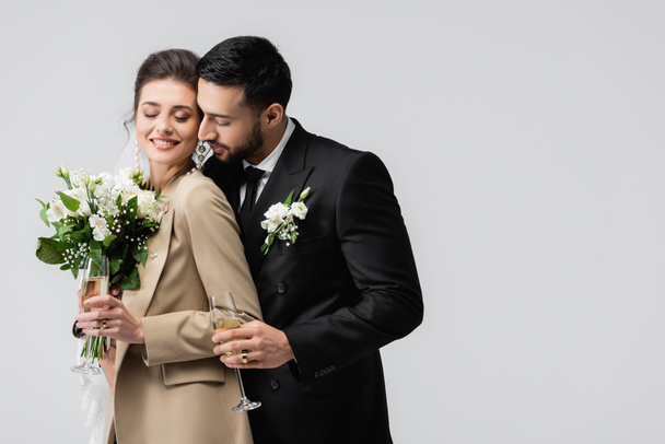 elegante árabe hombre con champán vidrio abrazo feliz novia celebración boda ramo aislado en gris - Foto, Imagen
