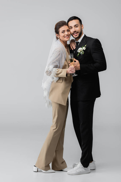 vista completa de los elegantes recién casados interracial de pie con copas de champán en gris - Foto, Imagen