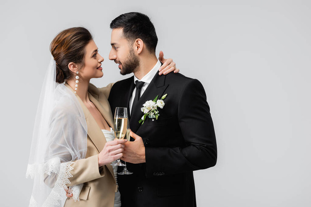 glückliche Braut mit Champagnerglas umarmt Hals von eleganten arabischen Mann isoliert auf grau - Foto, Bild