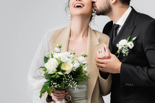 vue recadrée du marié arabe élégant embrassant mariée tenant bouquet de mariage et riant isolé sur gris - Photo, image