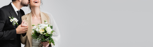 обрезанный вид мусульманина целующего взволнованную невесту держащую свадебный букет изолированный на сером, баннер - Фото, изображение