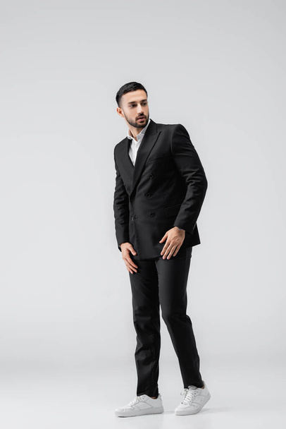 vue pleine longueur de jeune homme arabe en costume noir et baskets regardant loin tout en posant sur gris - Photo, image