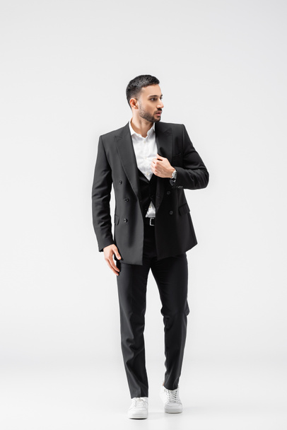 Ganzkörperansicht eines muslimischen Mannes in schwarzem Anzug und Turnschuhen, der wegschaut, während er auf grau läuft - Foto, Bild