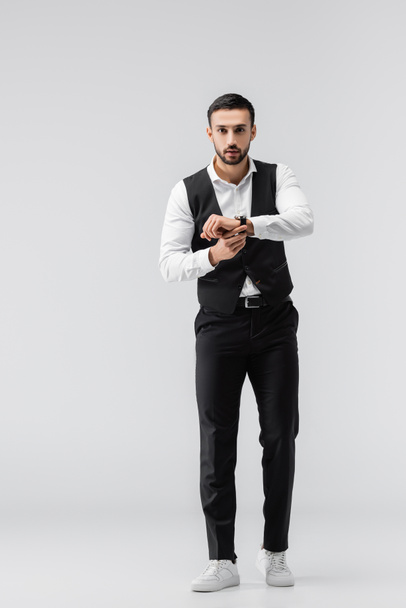 full length view of fashionable muslim man fixing wristwatch while looking at camera on grey - Photo, Image