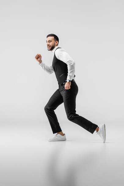 full length view of young arabian man in fashionable clothes and sneakers running on grey - Photo, Image