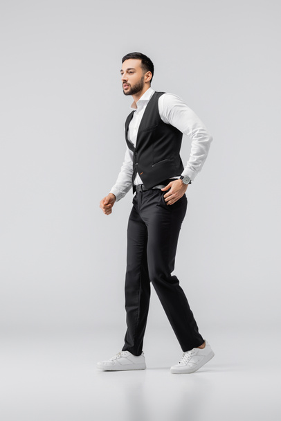 full length view of arabian man in elegant clothes and sneakers walking with hand in pocket on grey - Photo, Image