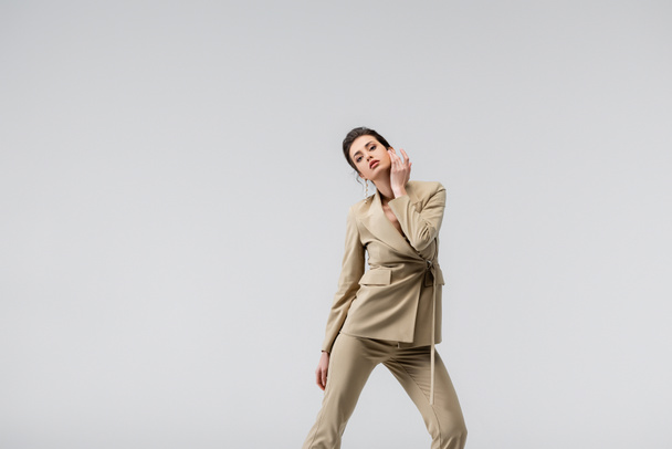 brunette vrouw in stijlvolle broekpak aanraken gezicht terwijl poseren geïsoleerd op grijs - Foto, afbeelding