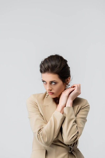 брюнетка женщина в стильном блейзере глядя в сторону, позируя с руками возле лица изолированы на сером - Фото, изображение