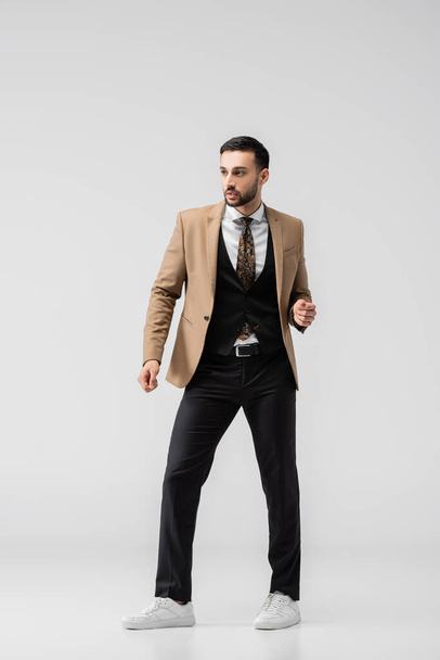full length άποψη του νεαρού muslim άνθρωπος στο μοντέρνο κοστούμι και sneakers κοιτάζοντας μακριά, ενώ θέτουν σε γκρι - Φωτογραφία, εικόνα