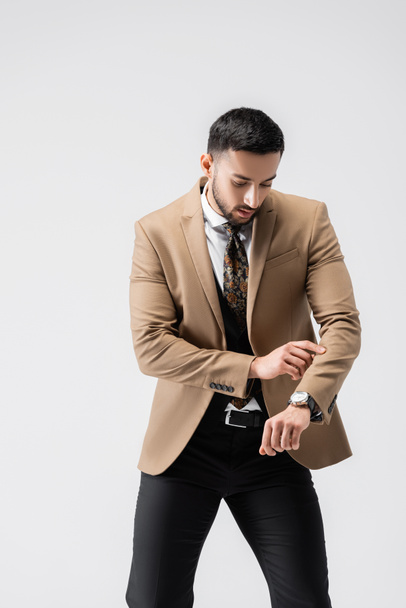 uomo arabo alla moda controllando il tempo sull'orologio da polso mentre posa isolato sul grigio - Foto, immagini