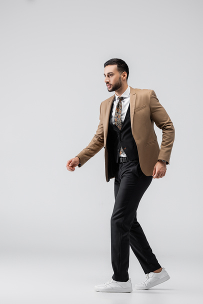 Ganzkörperansicht eines modischen muslimischen Mannes in Anzug und Turnschuhen, der auf grau läuft - Foto, Bild