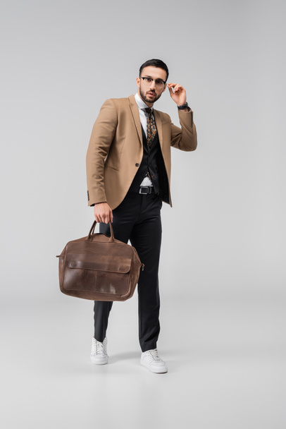 visão de comprimento total do homem árabe na moda tocando óculos enquanto posando com saco de couro no cinza - Foto, Imagem