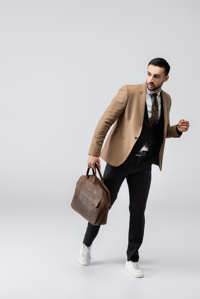 full length view of fashionable muslim man looking away while walking with leather bag on grey - Photo, Image