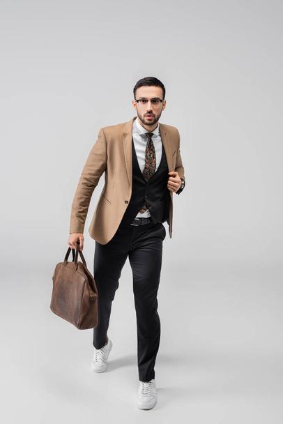 full length view of fashionable muslim man walking with leather bag and looking at camera on grey - Photo, Image