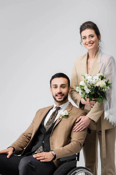 joyful woman with wedding bouquet touching shoulder of handicapped arabian fiance isolated on grey - Photo, Image