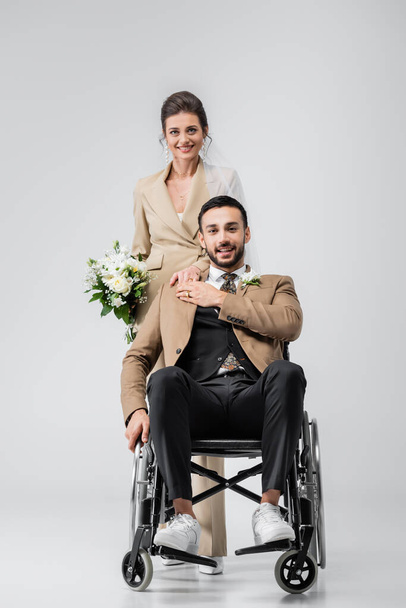 glücklicher, behinderter muslimischer Mann und elegante Braut mit Brautstrauß, die in die Kamera auf grau blickt - Foto, Bild