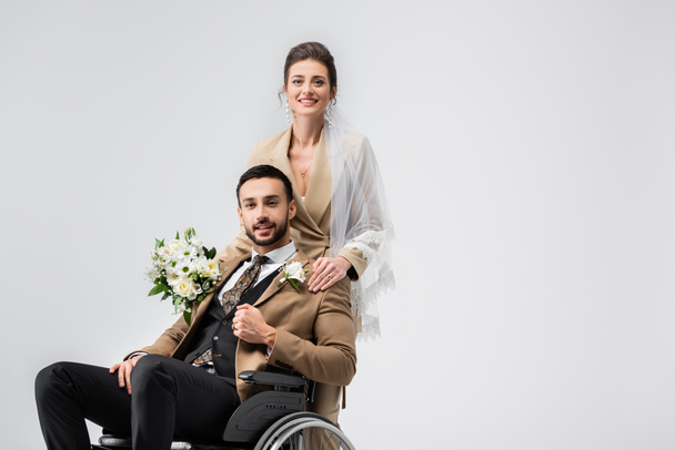 mujer elegante en velo, con ramo de bodas, mirando a la cámara cerca de musulmán discapacitado prometido aislado en gris - Foto, imagen