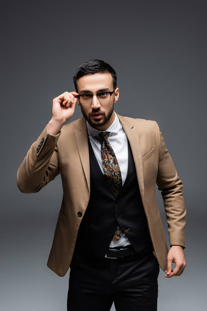 brunette muslim man in elegant suit, adjusting eyeglasses on grey - Photo, Image
